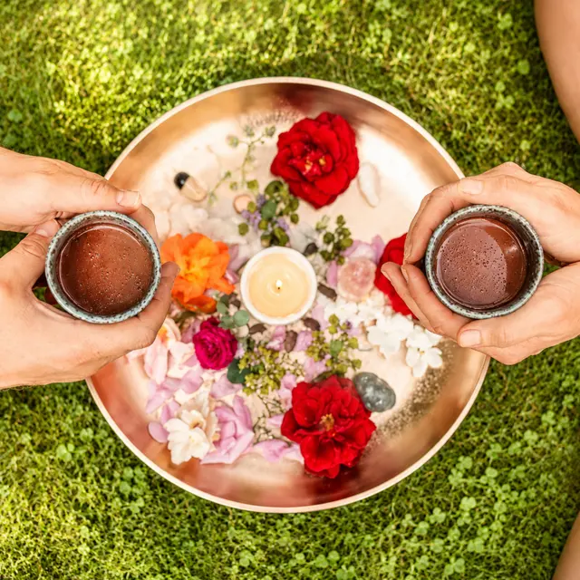 Zwei Personen halten Schalen mit heißem Kakaogetränk über einer dekorierten Schale mit Blumen und Kerze auf einer Wiese. Das Foto wurde mit Unterstützung künstlicher Intelligenz hinsichtlich Farben, Lichtstimmung und Bildqualität optimiert.